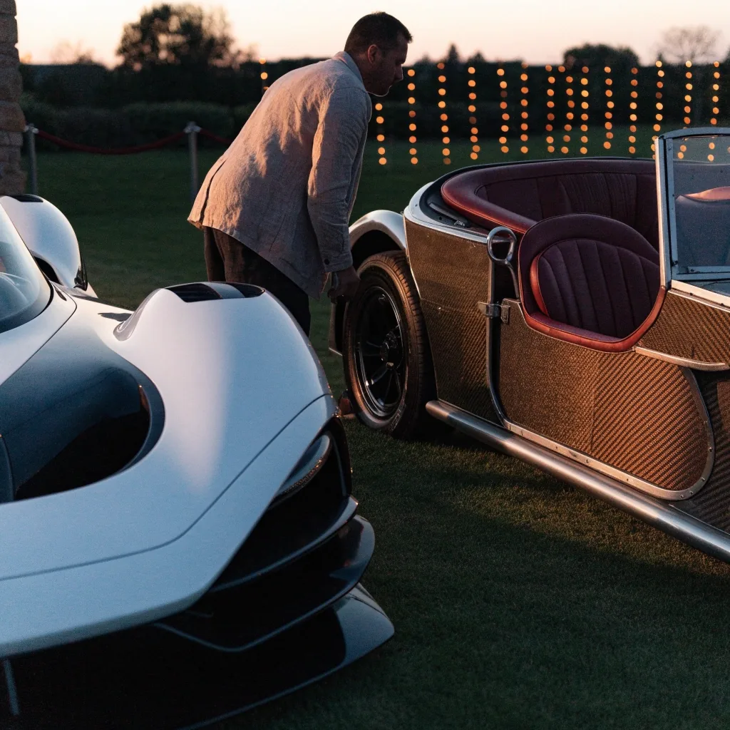Two unbadged hypercars of different generations displayed side by side at an evening gathering